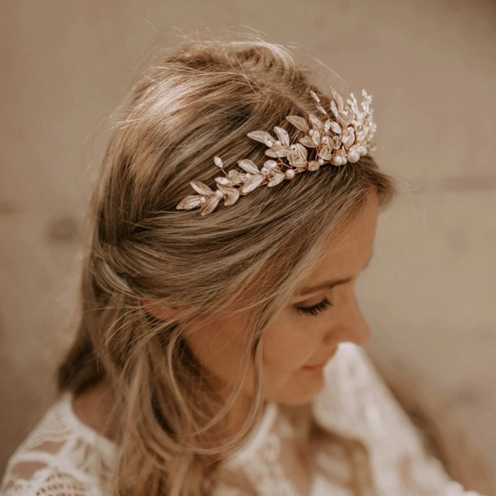 Ein zartes goldenes Diadem, verziert mit Metallblättern, kleinen Perlen und Kristallen, ruht auf einem Spitzenstoff neben einer hölzernen Oberfläche, die sanft in warmen Tönen beleuchtet ist.