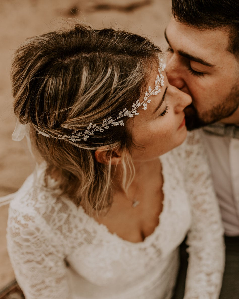 Eine Braut in weißem Spitzenkleid und geblümtem Braut Haarschmuck schließt sanft die Augen, während der Bräutigam mit Gesichtsbehaarung ihre Schläfe küsst. Der intime, warme Moment ist von oben aufgenommen und hebt die eleganten Brautaccessoires und die zärtliche Verbindung der beiden hervor.