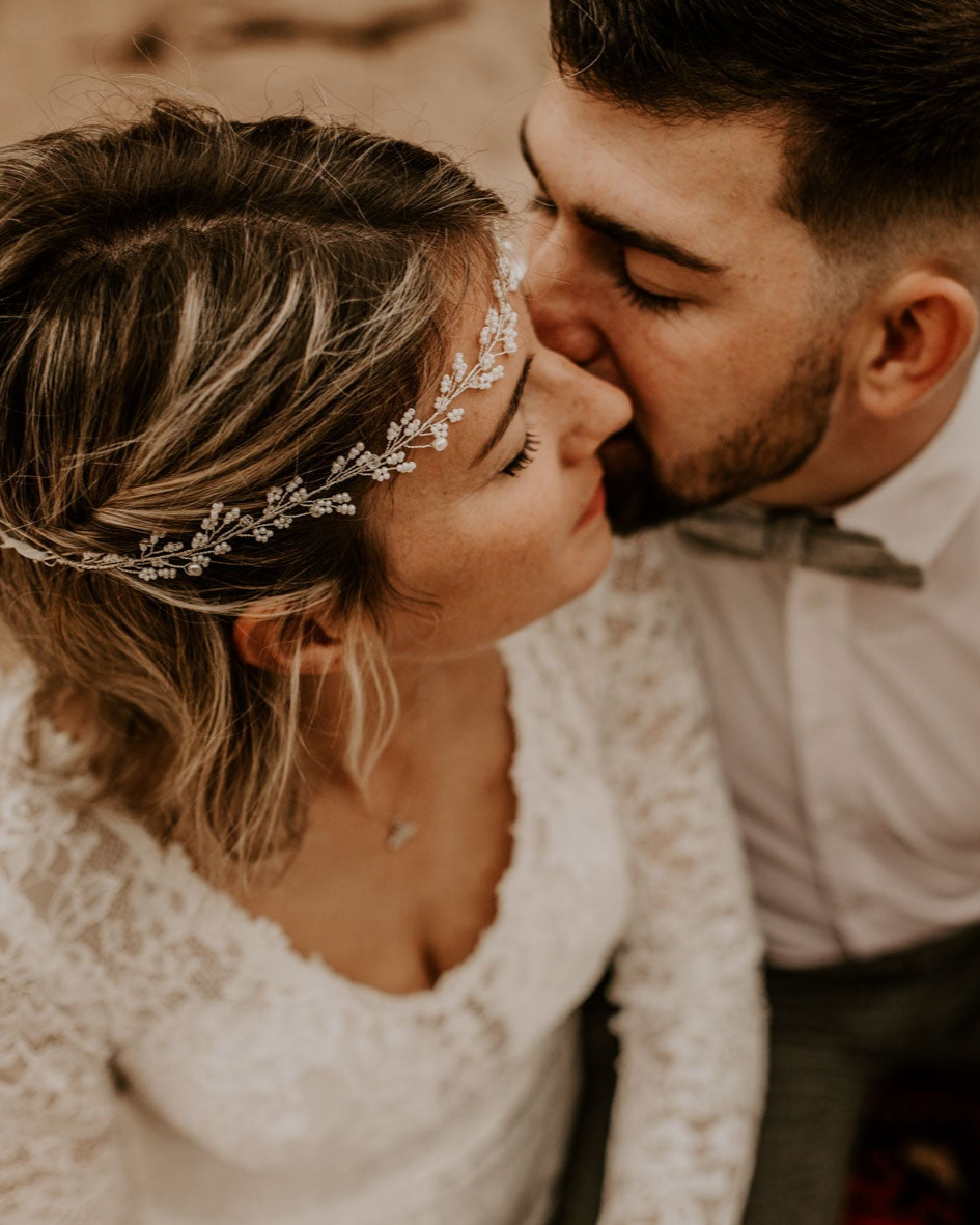 Eine Nahaufnahme von Braut und Bräutigam. Der Bräutigam, mit Gesichtsbehaarung, küsst sanft die Wange der Braut. Die Braut trägt ein Spitzenkleid und einen eleganten Haarschmuck Hochzeit in ihrem gewellten Haar. Beide wirken in ihrer formellen Hochzeitskleidung gelassen und intim.