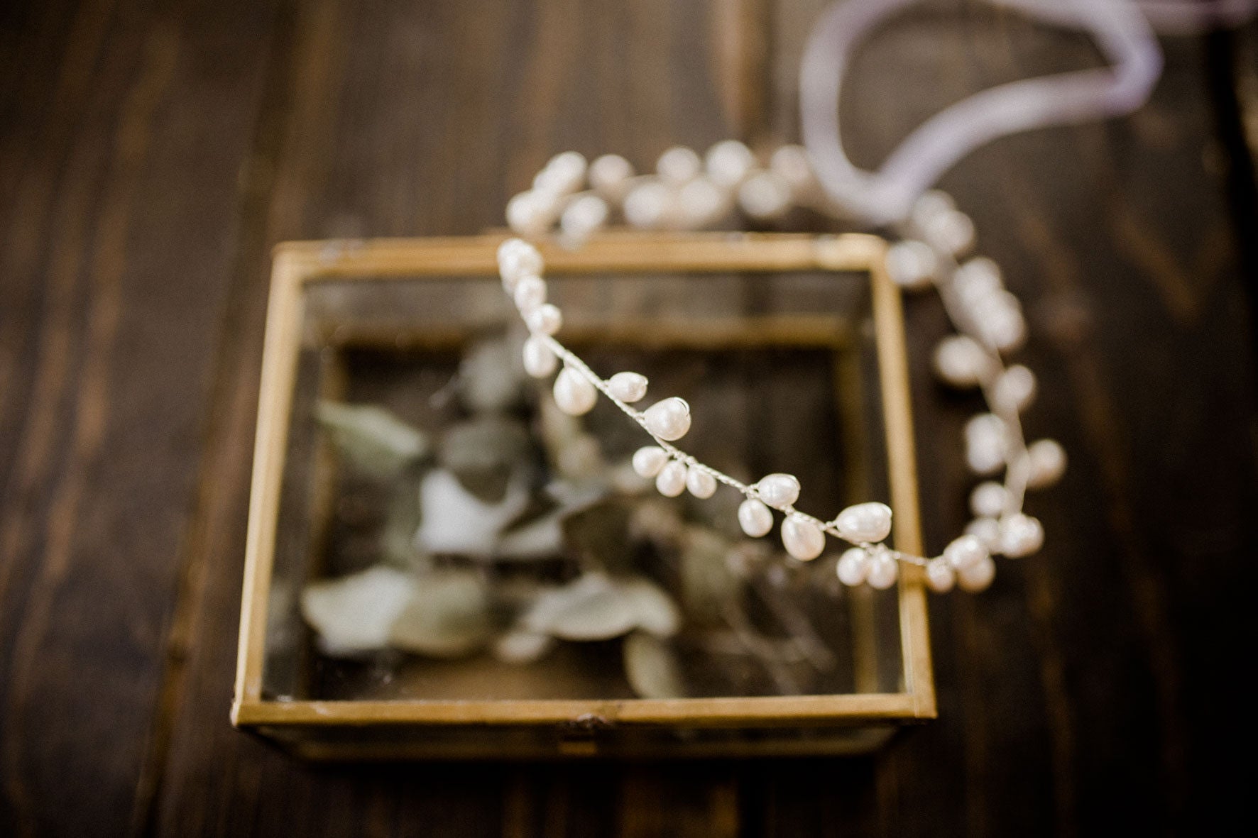 Ein zartes, mit kleinen weißen Perlen besetztes Braut Haarschmuck Stirnband ruht auf einem Schmuckkästchen aus Glas und Messing. Auf einer rustikalen Holzfläche mit zartem Grün im Inneren platziert, entsteht ein eleganter Vintage-Look, der sich perfekt für Brautaccessoires eignet.